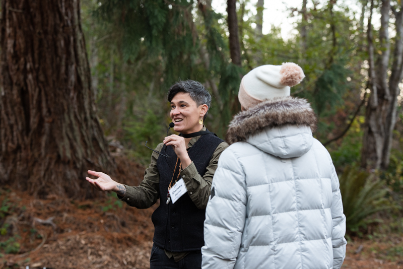 A person wearing a black vest with a green long sleeve is speaking into a microphone while a person in a grey jacket and multi-colored cap is standing listening in a forest.