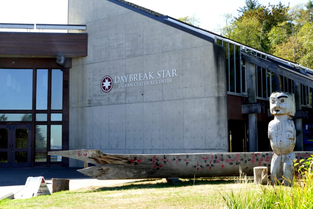 A concrete side of a building with "Daybreak Star Indian Cultural Center" on it. The building is behind a canoe and statue.