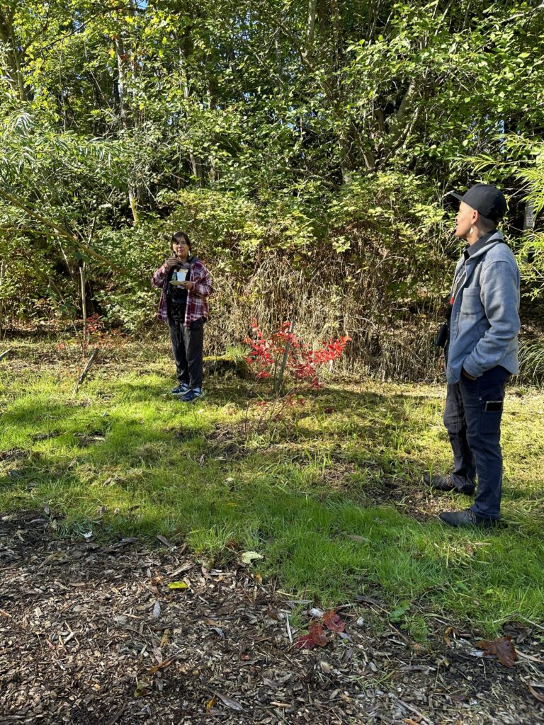 Two people, one in a red flannel and the other in a grey jacket standing next to a plant and explain the uses to a group.