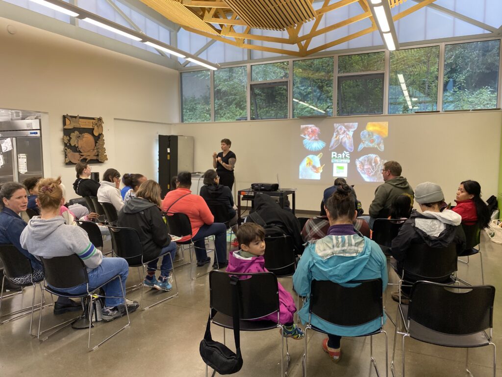 Person speaking at the Bat Night event to an audience of people in a large room, with a presentation of bats behind them.