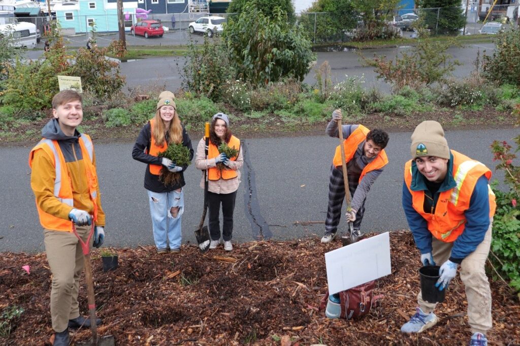 A group of five volunteers in bright orange vests smiling while planting trees along the trailside.