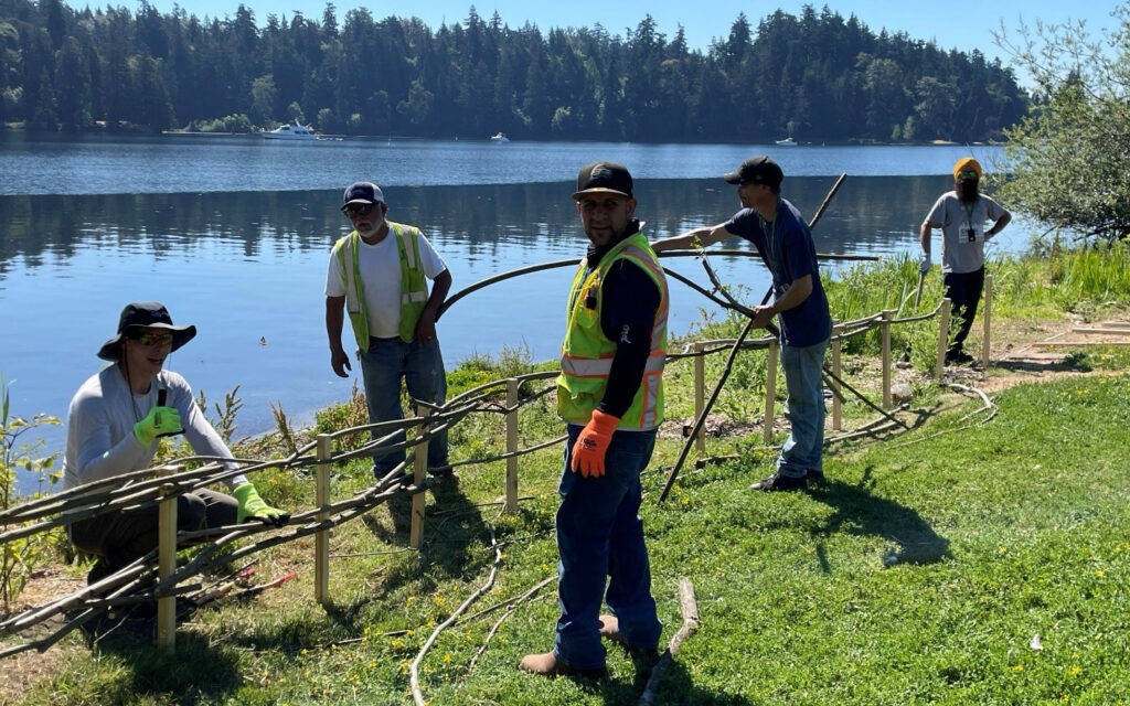 5 Natural area crew members working on building a fence with wooden sticks next to a lake.