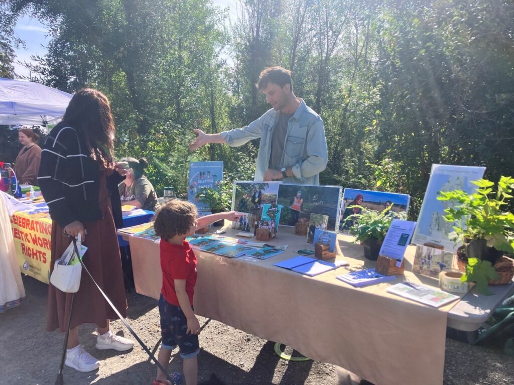 Michael standing behind a booth that is filled with photos, flyers, and plants, talking to a family