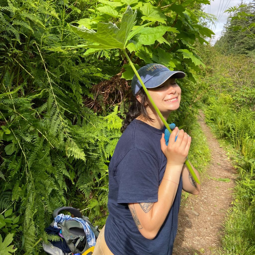 Cameryn standing among greenery holding a large tropical leaf like an umbrella.