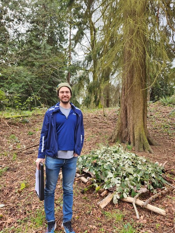 Greg standing in front of a platform of branches on the ground, with green leaves piled on top of it.